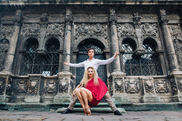 Fototapeta na wymiar beautiful happy couple in love embracing and dancing at old building in sunny street. stylish hipster groom and blonde bride in red dress performing dance, professional dancers. romantic moments