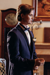 Obraz premium stylish groom in bow tie and blue suit getting ready in the morning in hotel, looking in mirror. wedding day concept. handsome man preparations in vintage room, in tuxedo with boutonniere