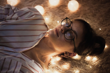 Awesome cute girl in white shirt and transparent glasses lying on the soft bed with fairy lights at night
