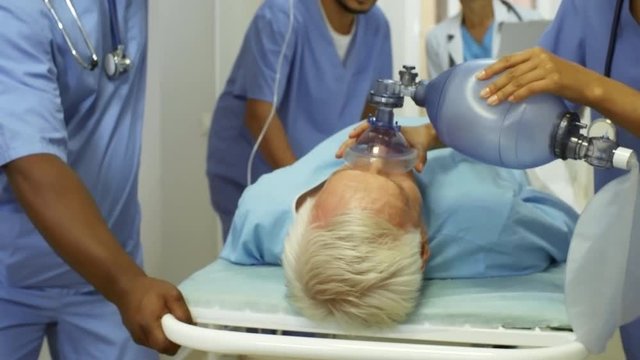 Dolly shot of multi-ethnic group of nurses in scrubs administering oxygen and IV fluids to elderly patient and rushing him to surgery as female doctor in lab coat giving instructions