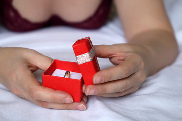 Woman opens gift box with golden ring, romantic present, marriage proposal. Seductive girl with sexy breast in red bra lies in bed, concept of jewelry, birthday, Valentines day