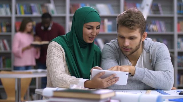 Beautiful muslim female in hijab and caucasian male students sharing digital tablet, discussing data online during case study in university library with blurry diverse students studying in background.