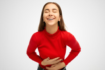 Positive and joyful young girl keeps both hands on belly, being in good mood, cant stopping laughing after hearing funny anecdote, isolated over white background in studio.