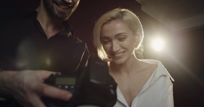 Close up of the male photographer showing photos of photoshoot to the young blond female model on the little camera screen in the dark studio.