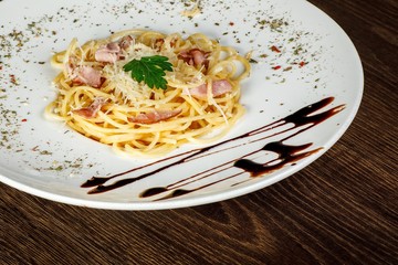 Fresh Italian pasta with bacon, cheese and green leaf in a big white dish decorated by sauce on a brown wooden table decorated by parmesan