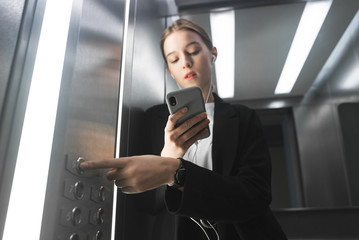 Female in the suit pushing the button it the elevator using gadgets. Close-up photo of an office worker, pushing the lift's button, using smartphone and headphones. © bodnarphoto