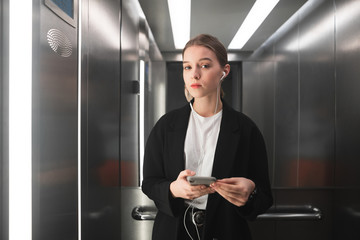 Young businesswoman is seriously looking at the camera in elevator using her smartphone and...