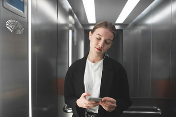 Attractive businesswoman is listening to music on her headphones with closed eyes in the elevator holding her smartphone. Employee with closed eyes is enjoying music in the lift using phone.