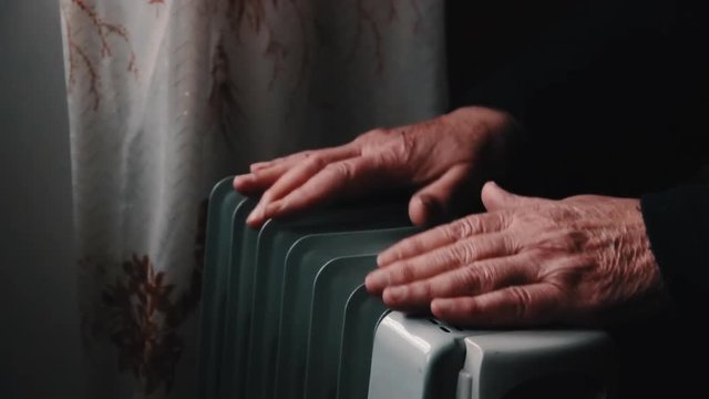 An elderly man warms his hands over an electric heater. In the off-season, central heating is delayed, so people have to buy additional heaters to keep houses warm despite increased electricity bills.