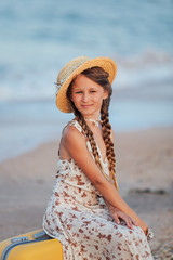 A little girl sits on a yellow suitcase on the beach. Portrait of a girl with a hat. Retro style