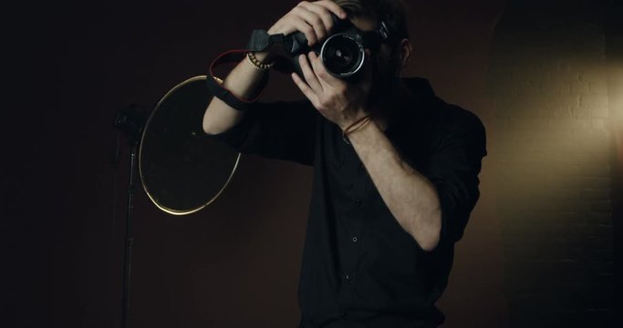 Caucasian male stylish photographer in the black shirt taking a photo with a camera in the dark studio amd smiling. Portrait shot.