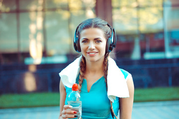 Young happy smiling woman is ready for outdoor workout, holding bottle of water, towel on neck and...
