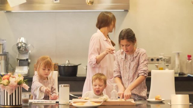 The mother talks with her children and checks how her daughter does dough circles for the future cookies. After she takes one circle and puts it on the pan at the back of her.