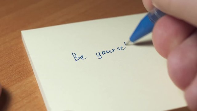 Someone is writing BE YOURSELF on a sheet of sticker paper. Close up shot, selective focus. Self motivation,personal growth