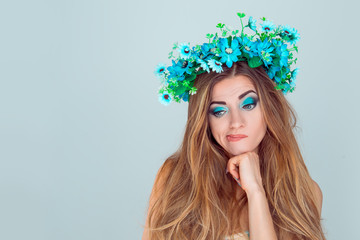 Head shot thoughtful puzzled upset young Woman with floral headband looking down to the side. Fashion girl with crown from sunflowers on head doubtful isolated on light blue background with copyspace