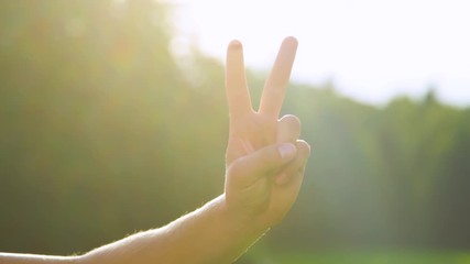 SLOW MOTION, CLOSE UP, LENS FLARE, DOF: Bright golden sunbeams shine on male hand and showing the peace sign. Unrecognizable young man shows the victory sign to the camera on a sunny summer day.
