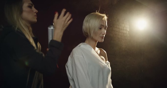 Hairdresser doing hair of the pretty blond female model during her photoshoot in the dark studio with a single lamp.
