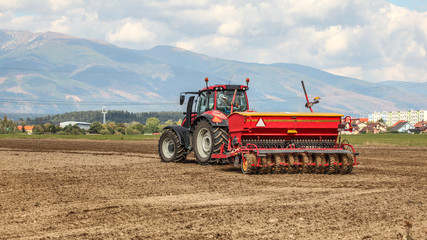 Fototapeta premium Red tractor pulling sowing trailer behind over empty field. Some village and mountains in background.