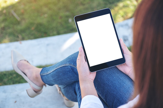 Mockup image of a woman holding and using black tablet pc with blank white desktop screen while sitting in the outdoors