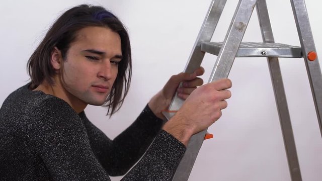 Handsome man in short black shiny gress coming up the stairs. Transgender man with hairy legs in stockings defiantly looking at the camera, smiling