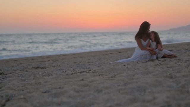 happy mom with her little daughter sitting on the beach at sunset, hugging and playing. concept of happy mothers and daughters