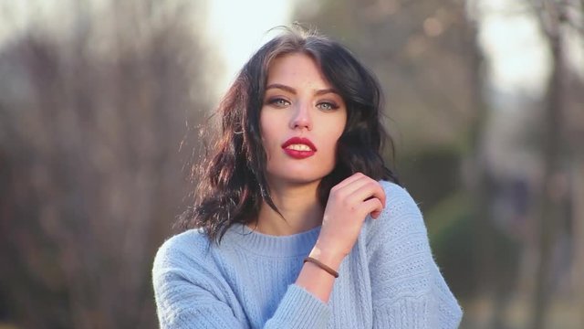 Close-up attractive sexy brunette girl with beautiful hair and red lips poses on camera depicting different emotions standing outdoors with blurred background. Portrait of young woman model.