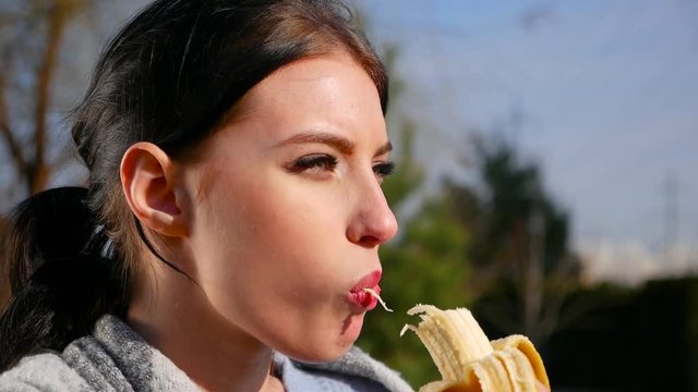 Slow motion, close-up attractive sexy young brunette woman with beautiful eyes and red lips eats banana on sunny day. Spring beriberi, vitamins in fruits.