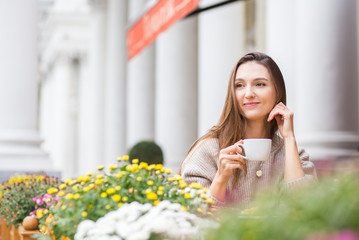 Young attractive smiling woman in gray knitted sweater drinking tea outdoors at the street cafe.