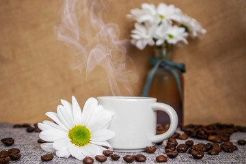 A cup of espresso with steam, next to it is a white flower, in the background a vase with flowers, a burlap background