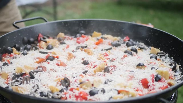 Close up of a paella being cooked and rice being pored on top of it with spices.