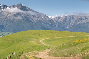 Fototapeta na wymiar Countryside landscape with dirt winding road among green meadows and snow capped Caucasus mountains in the background on a sunny summer day. Karachay-Cherkessia, Russia