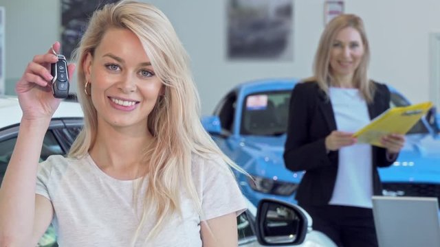 Gorgeous happy woman smiling, holding car keys after buying new auto. Attractive female driver buying new vehicle, professional car dealer on the background. Consumerism, rental concept