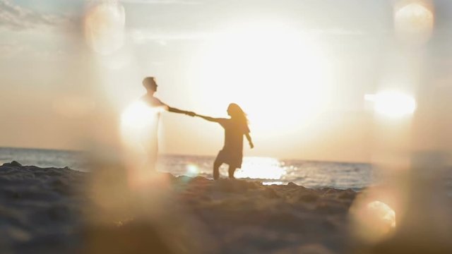 slow motion couple silhouettes dance on sandy beach against sea reflecting sun rays with blurred bottles on foreground