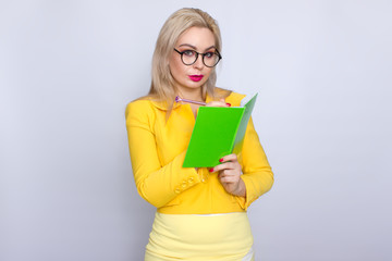 Close-up portrait of amazing blonde woman holding notebook, book, literature, in hands arms and pen pencil in yellow suit over white background in studio