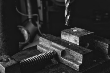 Black and white photo of drill bit coming down onto a piece of wood held in a vice in an industrial...