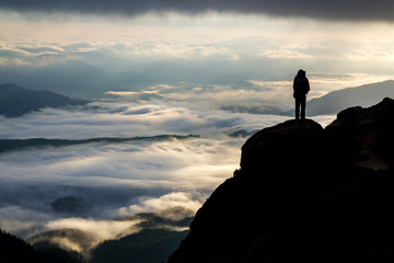 Wide mountain panorama. Small silhouette of tourist with backpack on rocky mountain slope over valley covered with white puffy clouds. Beauty of nature, tourism and traveling concept