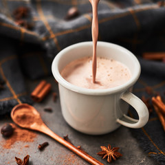 Pouring tasty hot cocoa drink into retro mug with ingredients: cinnamon, anis and cove on black concrete table. Winter drink. Copy space for text. Square crop.