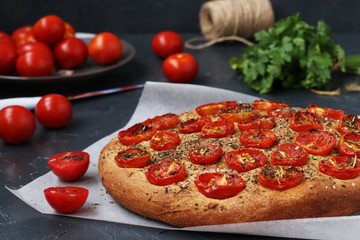 Focaccia with cherry tomatoes is located on parchment on a dark background. In the background is a bunch of parsley and a plate of tomatoes. Horizontal photo