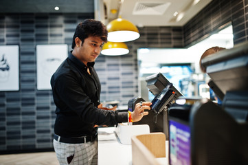 Indian man holding pay by credit card at cash desk with order screen and payment terminal in food...