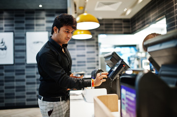Indian man holding pay by credit card at cash desk with order screen and payment terminal in food cafe.