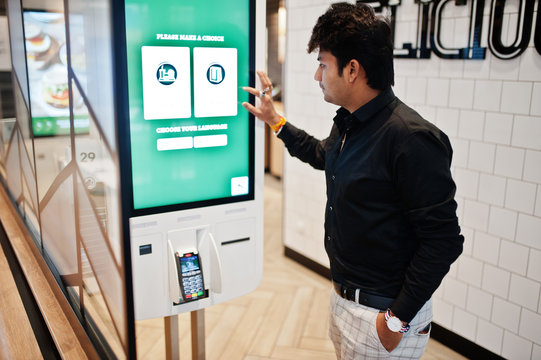 Indian man customer at store place orders and pay through self pay floor kiosk for fast food, payment terminal. Make a choise of language on screen.