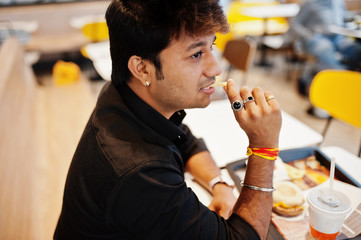Stylish indian man sitting at fast food cafe and eating french fries.