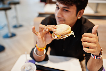 Stylish indian man sitting at fast food cafe and eating hamburger. Show thumb up.
