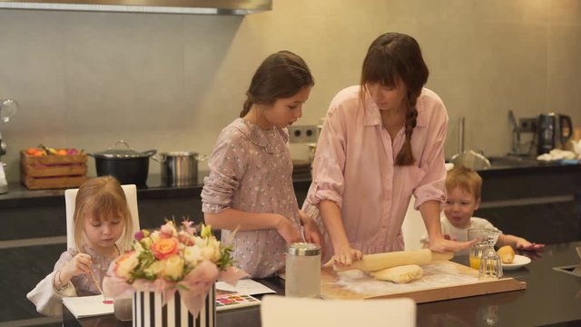 The mother teaches her daughter step by step how to roll the dough using rolling pin. So, the girl can do it herself after. Other kids are busy at the table.