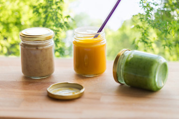 baby food, healthy eating and nutrition concept - vegetable or fruit puree in glass jars and feeding spoon on wooden table over green natural background