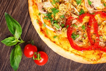 Top view close up Italian pizza with melted cheese, red tomatoes, pepper, mushrooms, onion and herbs on a brown table decorated by red cherry tomatoes and fresh green basil leaves