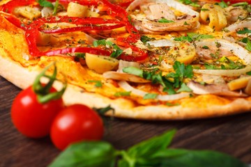 Close up Italian pizza with melted cheese, red tomatoes, pepper, mushrooms, onion and herbs on a brown table decorated by cherry tomatoes  and fresh green basil leaves