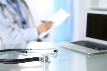 Stethoscope lying on glass desk with laptop computer at busy physician background. Medicine or pharmacy concept. Medical tools at doctor working table