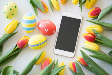 easter, holidays, tradition and object concept - smartphone with colored eggs and tulip flowers on...