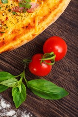 Top view close up mushrooms and Italian pizza with ham, cheese, pepper and herbs on a brown table decorated by fresh red cherry tomatoes and fresh green basil leaves